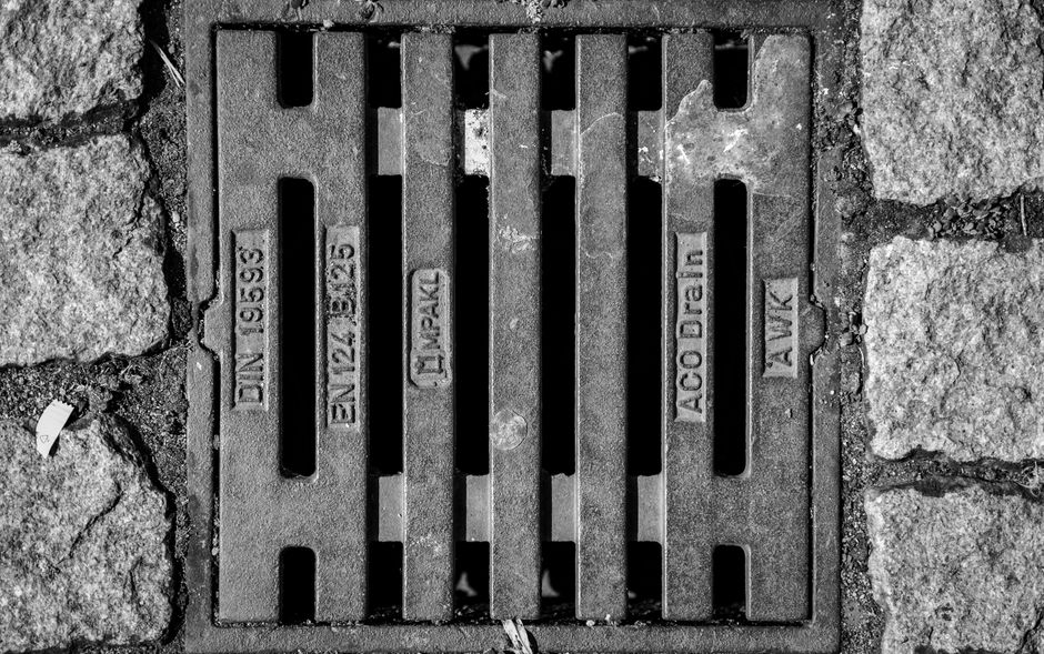A detailed close-up of a street drainage grate flanked by cobblestones in black and white.